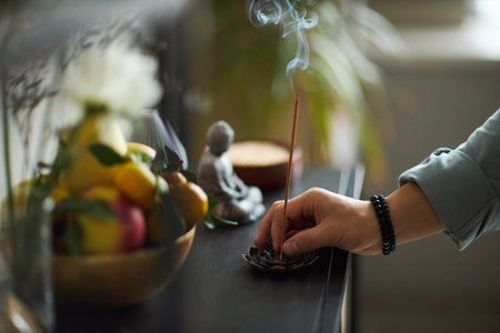 Asian young adult hand placing incense stick into holder with smoke rising, fruit bowl and small Buddha statue visible in background, indoor setting implied by contextの写真素材