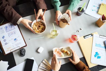 Young Asian businessmen and businesswomen sitting at office table eating lunch with chopsticks, discussing documents and charts, hands visible holding food containers and drinks during workの写真素材