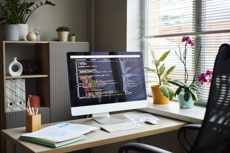 Young Asian man working at desk using desktop computer with programming code on screen, surrounded...