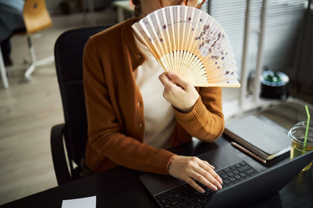 Young adult Asian woman sitting at desk using laptop, holding decorative hand fan in front of face,...