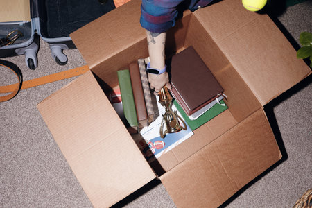 Caucasian girl hand with tattoo and smartwatch packing books, notebooks, and trophy into cardboard box on carpeted floor, suitcase and tennis ball partially visible in backgroundの写真素材