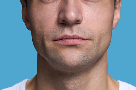 Caucasian young adult man showing lower half of face with neutral expression against blue background, lips closed, stubble visible, wearing casual shirtの写真素材