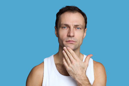 Portrait of Caucasian middle aged man touching chin with hand while looking into camera, showing thoughtful facial expression against plain blue background, wearing sleeveless shirtの写真素材