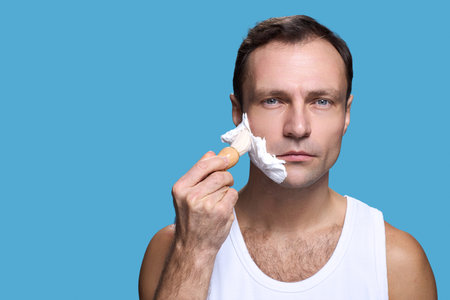 Portrait of middle aged Caucasian man looking into camera applying shaving foam to face with shaving brush against blue background, wearing sleeveless undershirt, showing serious expressionの写真素材