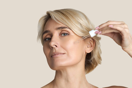 Portrait of middle aged woman applying facial serum with dropper to cheek, showing skincare routine...