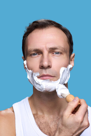 Portrait of Caucasian middle aged man looking into camera applying shaving cream to face with shaving brush against blue background showing daily grooming routineの写真素材