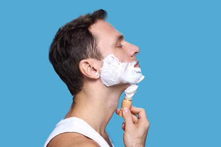 Caucasian young adult man applying shaving cream to face with shaving brush, eyes closed, side profile against blue background, demonstrating personal grooming routineの写真素材