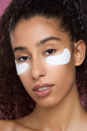 Portrait of young adult woman looking at camera, wearing under eye patches on face showing skincare routine against plain background with natural makeup and curly hair visibleの写真素材