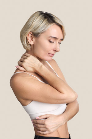 Caucasian middle aged woman touching neck and shoulder with closed eyes, standing sideways against...
