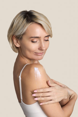 Caucasian middle aged woman applying moisturizing cream to shoulder, smiling with eyes closed, showing healthy skin and self care routine, wearing sleeveless top, neutral backgroundの写真素材