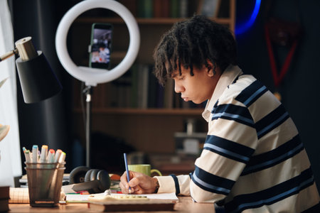 Teenage boy studying at desk, writing in notebook with pen, focusing on homework while smartphone...