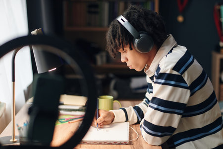 Teenager boy wearing headphones sitting at desk studying and writing in notebook, focused on...