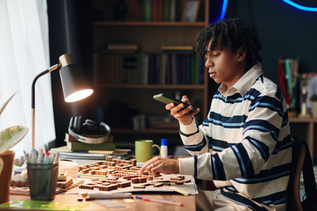 Teenage Black boy sitting at desk using smartphone while assembling wooden puzzle or architecture...