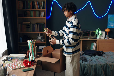 Black teenage boy unpacking trophies from cardboard box in bedroom, standing near desk with various...