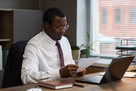 Middle aged Black man sitting at desk reading document, wearing glasses and formal attire, working...