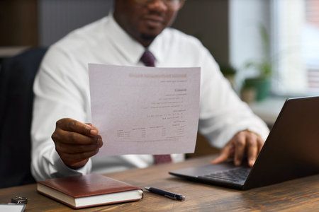 Middle aged Black man sitting at desk holding document toward camera while working on laptop, likely...