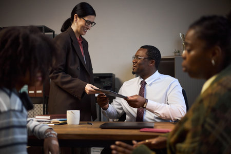 Middle aged Caucasian woman standing and handing documents to seated middle aged Black man in office...