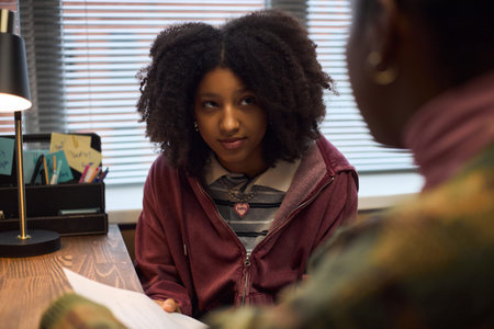 Teenage Black girl sitting at desk listening attentively to middle aged Black woman holding document...