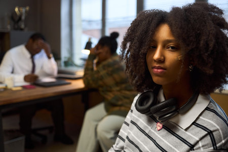 Portrait of Black teenage girl with headphones around neck sitting in office with serious...