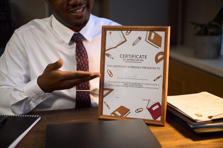 Middle aged man sitting at desk smiling and pointing at framed certificate of appreciation,...