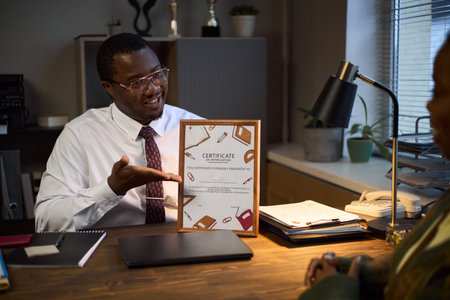 Middle aged Black man wearing glasses sitting at desk smiling and presenting certificate to unseen...