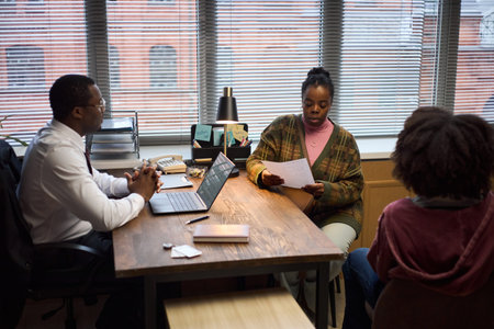 Middle aged Black man sitting at desk meeting with young Black woman holding documents and Black...