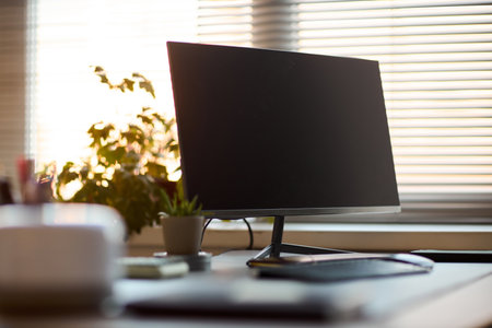 Modern computer monitor standing on office desk near keyboard and mouse with potted plant and blurred window blinds in background, technology workspace conceptの写真素材