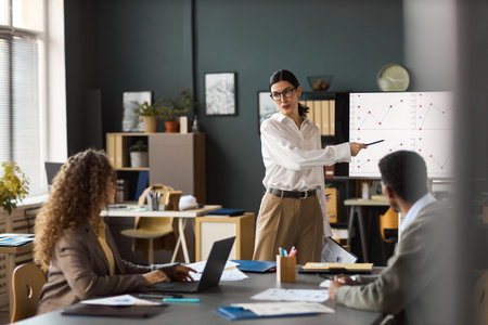 Caucasian young woman presenting data on screen to diverse colleagues in modern office, gesturing toward chart while explaining business statistics during meetingの写真素材