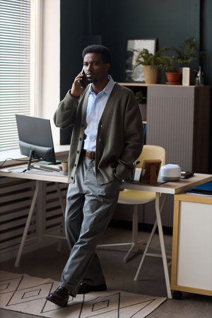 Serious Black man standing in modern office talking on smartphone, leaning against desk with computer monitor and office supplies near window blindsの写真素材
