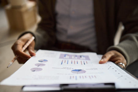 Middle aged Black man holding clipboard with financial charts, analyzing business data with pencil in hand, closeup of hands and documents, office setting visible in backgroundの写真素材