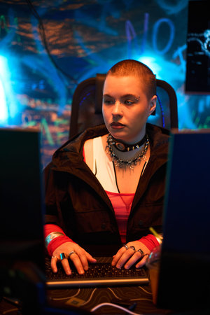 Young adult Caucasian woman with shaved head and facial piercings sitting at desk using computer, hands on keyboard, focused on screen, illuminated by neon blue lightsの写真素材