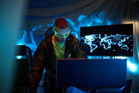 Young adult man wearing protective glasses operating multiple computer monitors in dark room, with...