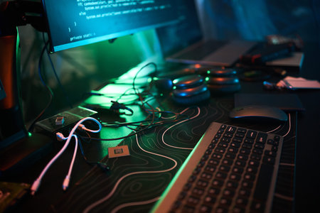 Closeup showing computer keyboard, mouse, circuit boards, headphones, and monitor displaying code,...