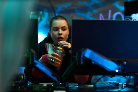 Young adult Caucasian woman sitting at workstation using desktop computers, holding plastic cup with straw, concentrating on screens in dark room with neon graffiti backgroundの写真素材