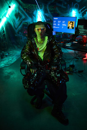 Young adult Caucasian man sitting in dark room wearing reflective jacket and futuristic glasses, working on a computer with multiple monitors displaying code and dataの写真素材