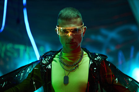 Portrait of Caucasian young adult man wearing futuristic glasses and layered chains standing in neon-lit environment, looking directly at camera with confident expressionの写真素材