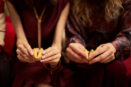 Young adult Asian woman and young adult Caucasian woman sitting side by side holding fortune cookies with both hands, preparing to break them open during holiday celebrationの写真素材