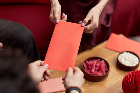 Asian woman handing red envelope to Asian young adult during Tet holiday celebration, hands visible...