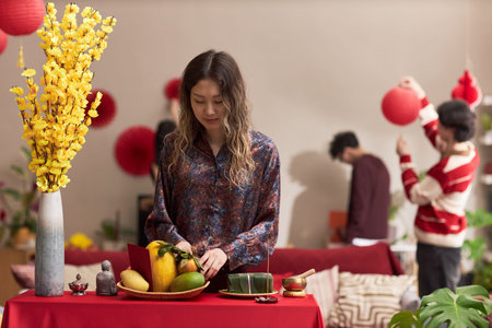 Young adult Asian woman arranging traditional Tet holiday fruits and cakes on table, standing...