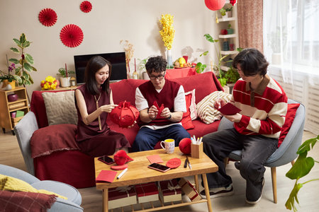 Three Asian young adults sitting on sofa preparing red Tet holiday decorations, holding paper crafts and yarn, engaging in traditional festive activity in living room settingの写真素材