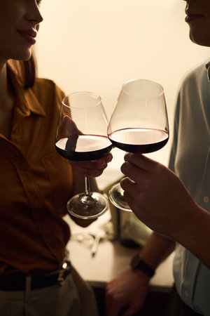 Caucasian young adult woman and Caucasian young adult man clinking wine glasses, standing close...