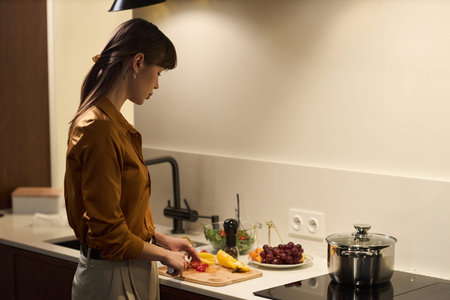 Young adult woman slicing vegetables on kitchen counter, preparing meal with fresh produce including...