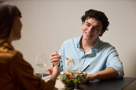 Young Caucasian man smiling while eating pasta and sitting across from young woman at table, holding...