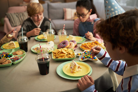 Group of Caucasian and multiethnic children wearing party hats sitting around table eating cake,...
