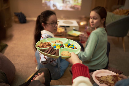 Two teenage girls sitting on floor turning heads toward camera while someone offering tray of...