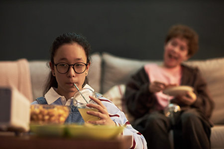 Asian teenage girl wearing glasses sitting on sofa drinking from plastic cup with straw while eating...