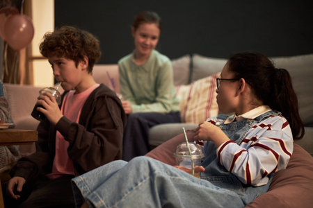 Three kids sitting together in living room, one boy drinking from plastic cup with straw while two girls talking and holding drinks, relaxed social interaction among childrenの写真素材