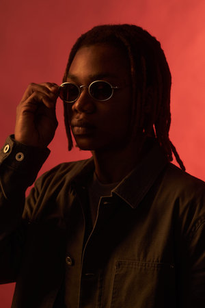 Young adult Black man adjusting sunglasses while facing camera, wearing casual jacket, standing against plain red background, showing confident expression, short dreadlocks visibleの写真素材
