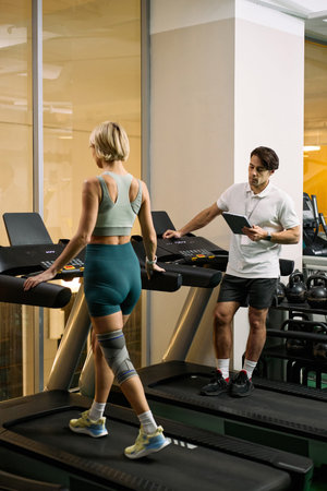 Caucasian young adult woman with knee brace walking on treadmill, under supervision of Caucasian...