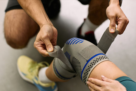 Caucasian young adult man assisting Caucasian young adult woman by wrapping elastic bandage around knee during physical therapy rehabilitation session focusing on injury recoveryの写真素材